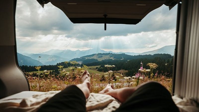 A picturesque mountain setting framed by the open doors of a camper van. A pair of reclining legs can be seen in the foreground.