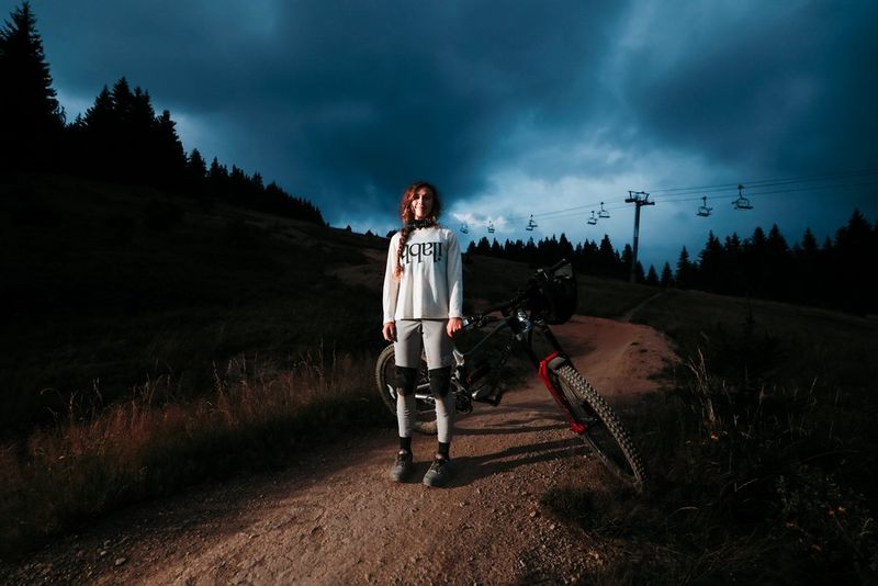 A woman stands on a dirt track holding up her mountain bike, a dark cloudy sky and silhouetted trees behind her. Taken on a Canon EOS R6 Mark II.