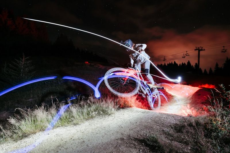 A mountain biker surrounded by red and blue light trails performs a jump against a dark sky. Taken on a Canon EOS R6 Mark II.