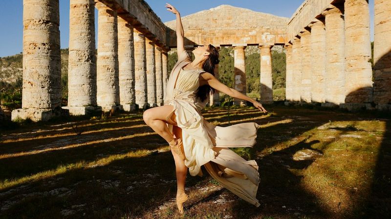A dancer wearing a flowing cream dress and ballet shoes poses en pointe amid the ruins of a Sicilian temple.