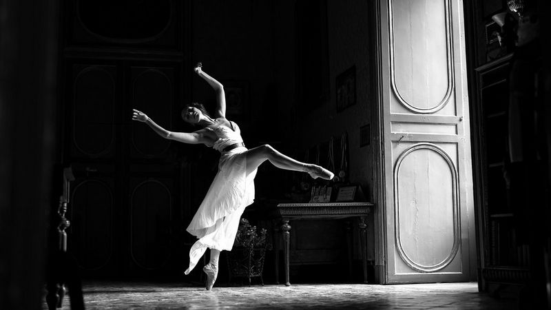 A black and white image of a ballet dancer in motion in a large, opulent room. Sunlight streams through an open door. 