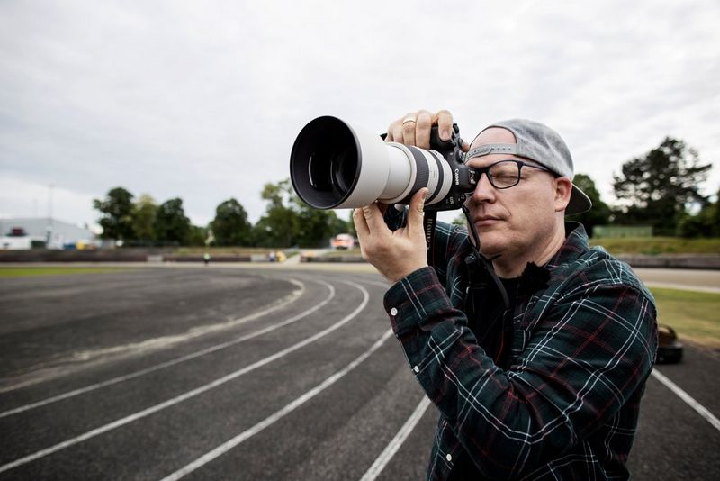 A man stands at the edge of a racetrack holding up a camera with a large super-telephoto lens to his eye.
