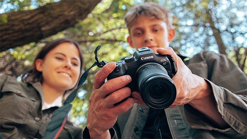 A Canon camera and lens taken from below, with two smiling young people slightly blurred in the background looking at the touchscreen. 