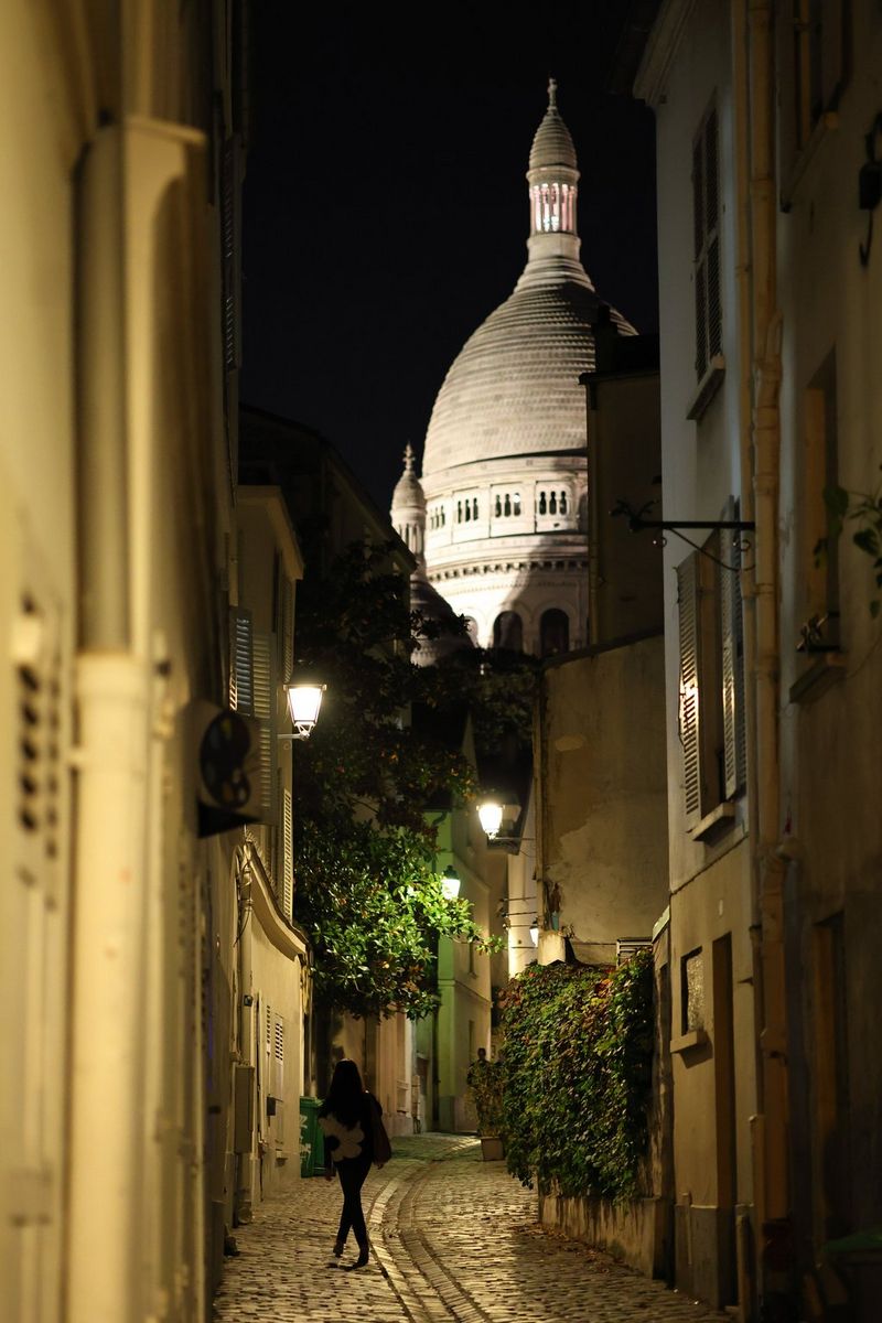 A woman walking on the cobbled Rue Saint-Rustique in Montmartre, at night, with the Sacré-Cœur Basilica visible in the distance, shot on a Canon EOS R8. 