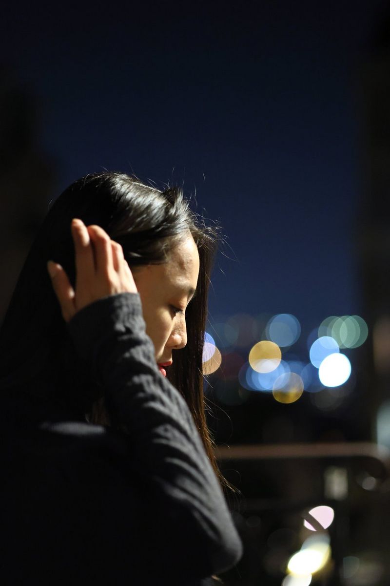 A woman photographed from the side using a Canon EOS R8 in low-light conditions, with out-of-focus lights blurred in the background. 