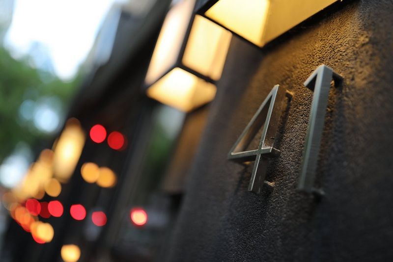 A close-up of the side of a building, a metal door number lit from the wall light above.