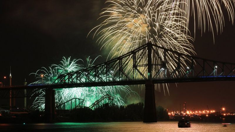 Multicoloured fireworks explode in the dark skies above a bridge, illuminating the water below. 