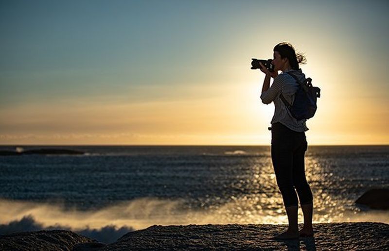 A person taking a photograph with a Canon camera and lens on a beach at sunset.