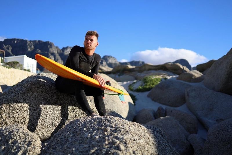 A man in a wetsuit sits on a rock on a beach, holding a surfboard, shot from a lower angle.
