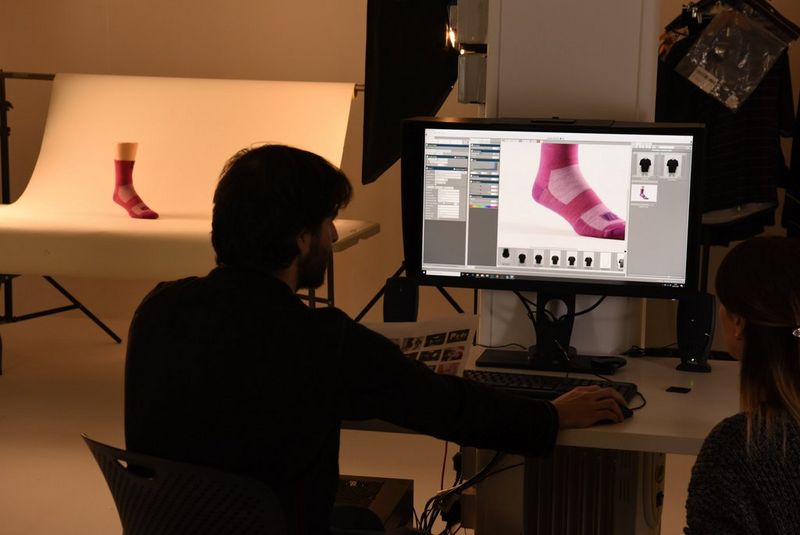 A man looking at a computer screen with an image of, and information about, a pink sock. The sock can be seen in the background, worn by a mannequin leg in a StyleShoots photography machine.
