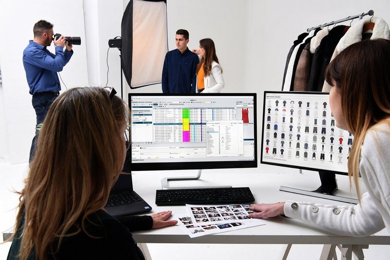 Two women looking at monitors, one with images of dozens of articles of clothing, the other with more detailed information on the clothing, while a photoshoot takes place in the background.