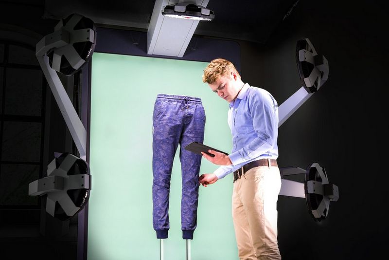A man looking at a tablet, next to a pair of purple trousers in a StyleShoots Vertical photography machine in front of a green backdrop.