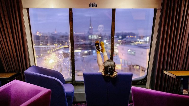 A girl sits in a purple armchair, surrounded by other purple armchairs. In front of her is a large dirty window that overlooks a city. She rests her socked feet against the window.