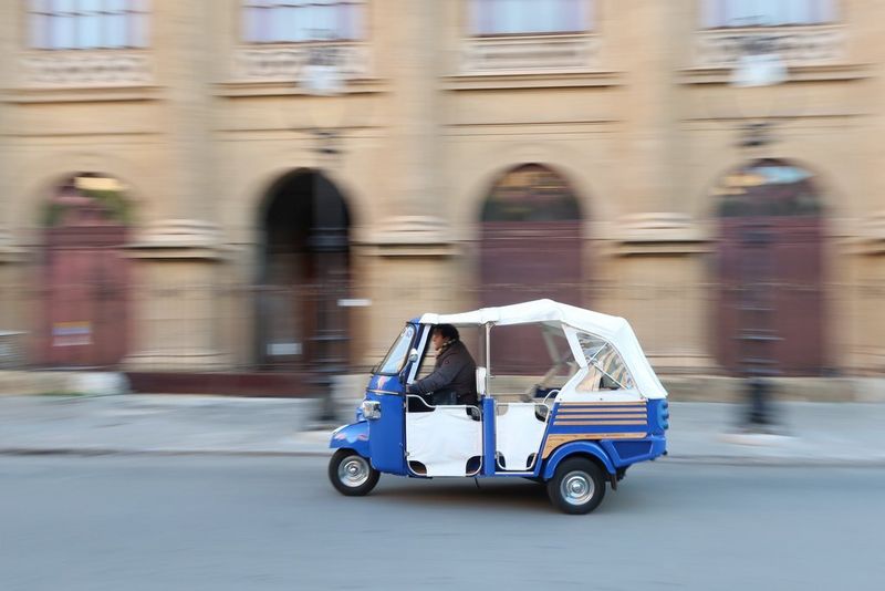 A small vehicle travelling along a road, passing buildings blurred because of camera panning.