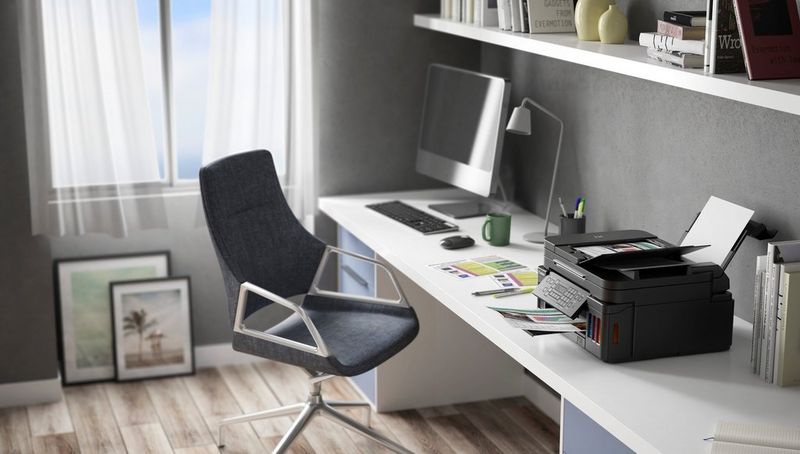 A Canon MegaTank printer sits on a desk in a home office, next to a computer and chair and near a large window.