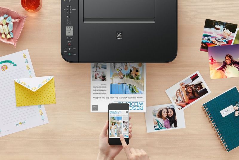 A Canon PIXMA printer, black in colour, prints a sheet of A4 paper while surrounded by pictures and printouts, and a hand holding a smartphone