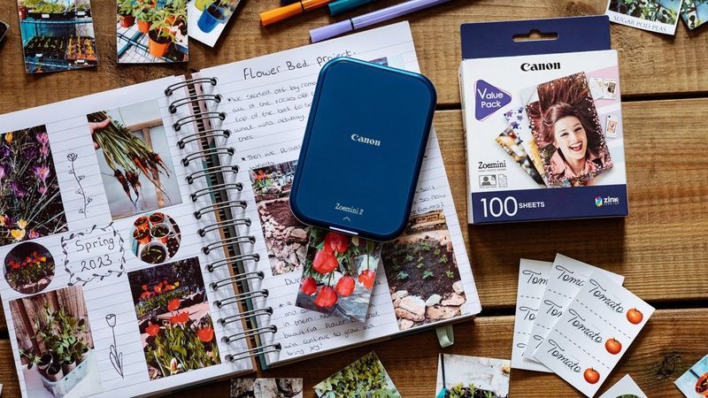 A print emerges from a Canon Zoemini 2 portable printer sitting on a journal, surrounded by stickers, prints, stationery and a 100-sheet pack of Canon ZINK™ 5 x 7.6cm Photo Paper. 
