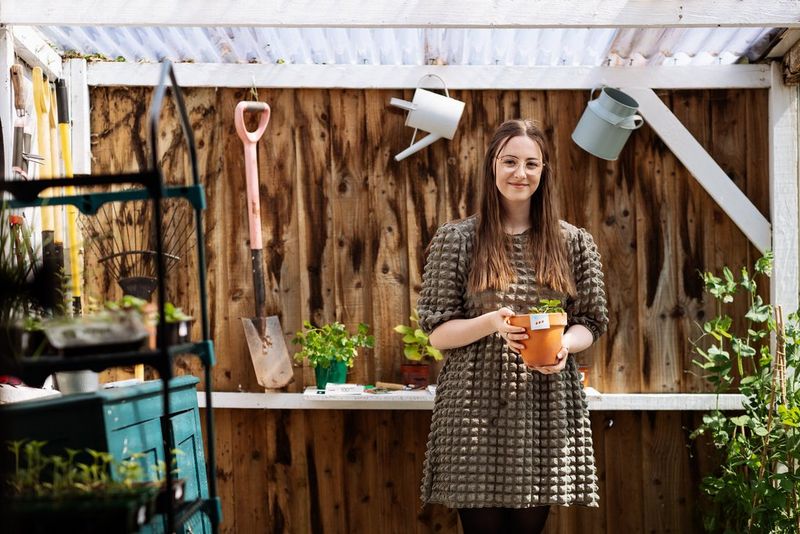 A young woman holding a plant stands in a potting shed, with a spade, a watering can and an urn hanging from the wall behind her.