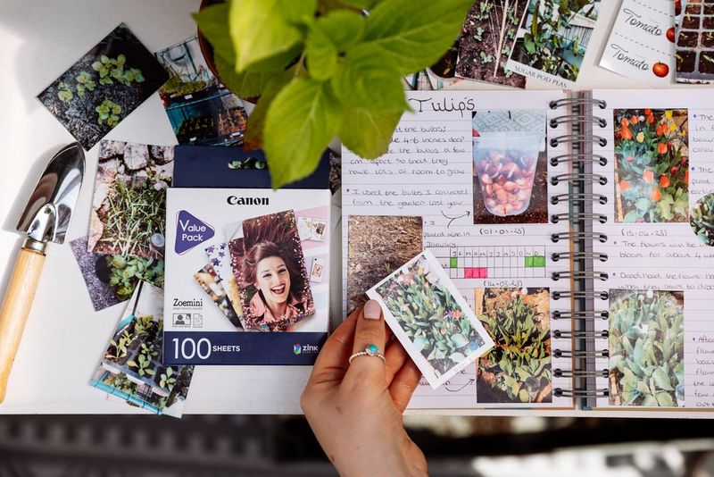A hand holding a print showing tulips in flower above a gardening journal. A 100-sheet pack of Canon ZINK 2 x 3in Photo Paper is on the desk beside the journal.