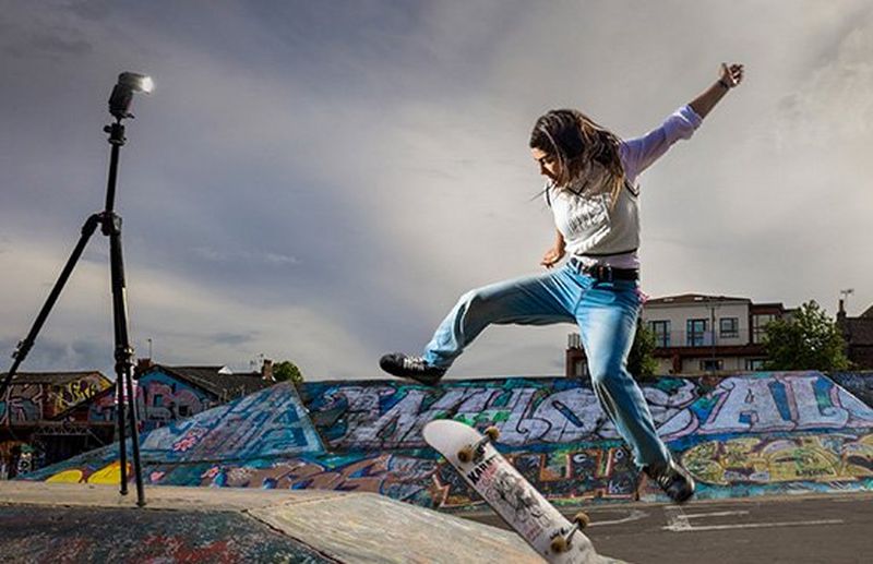 A woman skateboarding, captured frozen in mid-air by a Speedlite on a tripod, her board flipping beneath her.