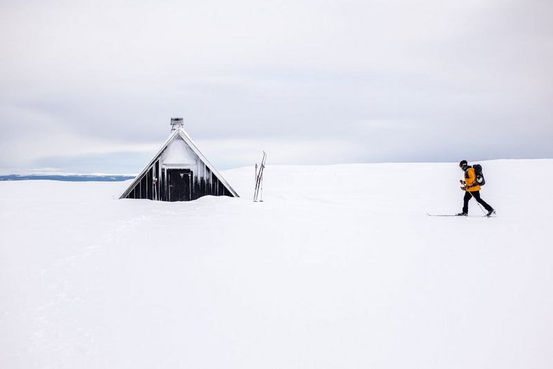 A man in a yellow jacket hikes across the snow towards a triangular chalet.