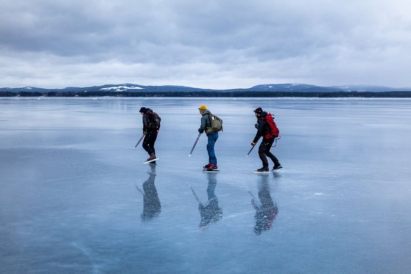Three skaters carrying backpacks traverse a vast frozen lake. Snow-covered peaks are visible in the distance.