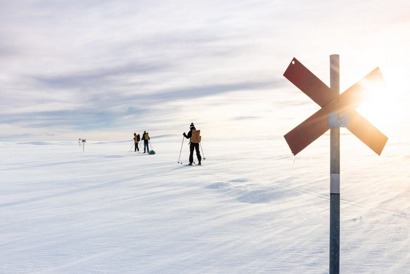 Three cross-country skiers traverse a snow-covered landscape. The sun's rays illuminate a marker pole behind them.