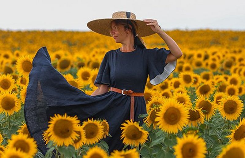 A woman in a long dress and a sun hat looks to the left as she stands in a sunflower field.