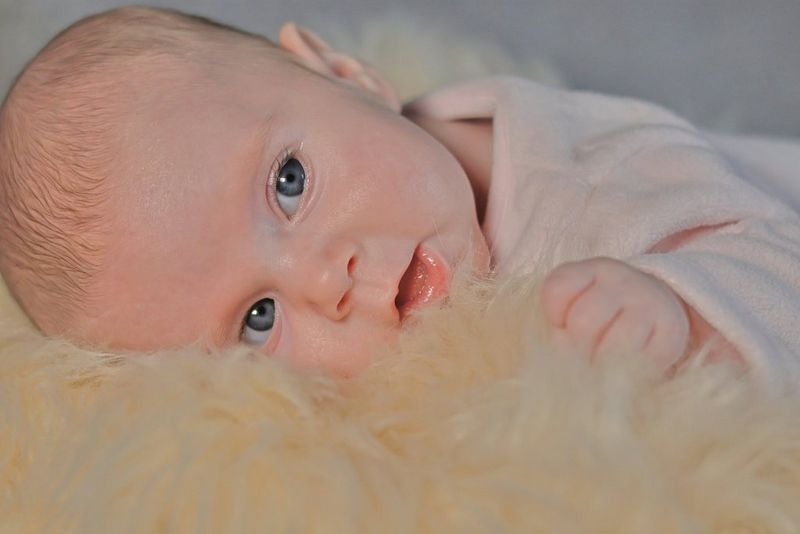 An infant wearing light pink top lying on the side on a fluffy beige blanket.
