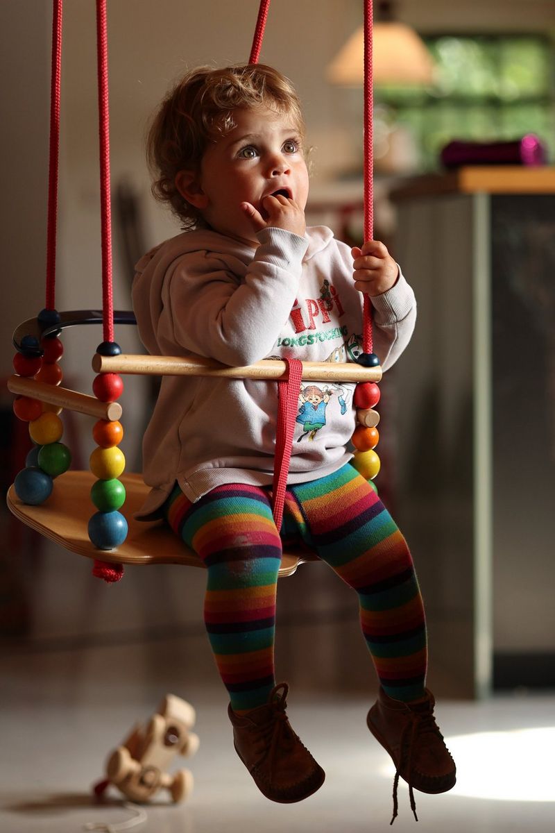 A toddler sitting in a swing chair looking at the window through which natural light is coming in.