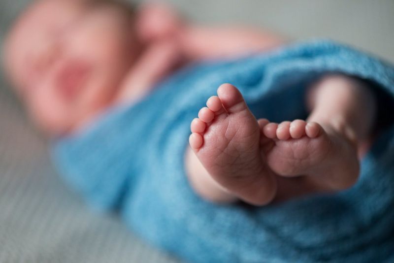An infant wrapped in a blue blanket lying on a bed with their little pink feet in focus. 
