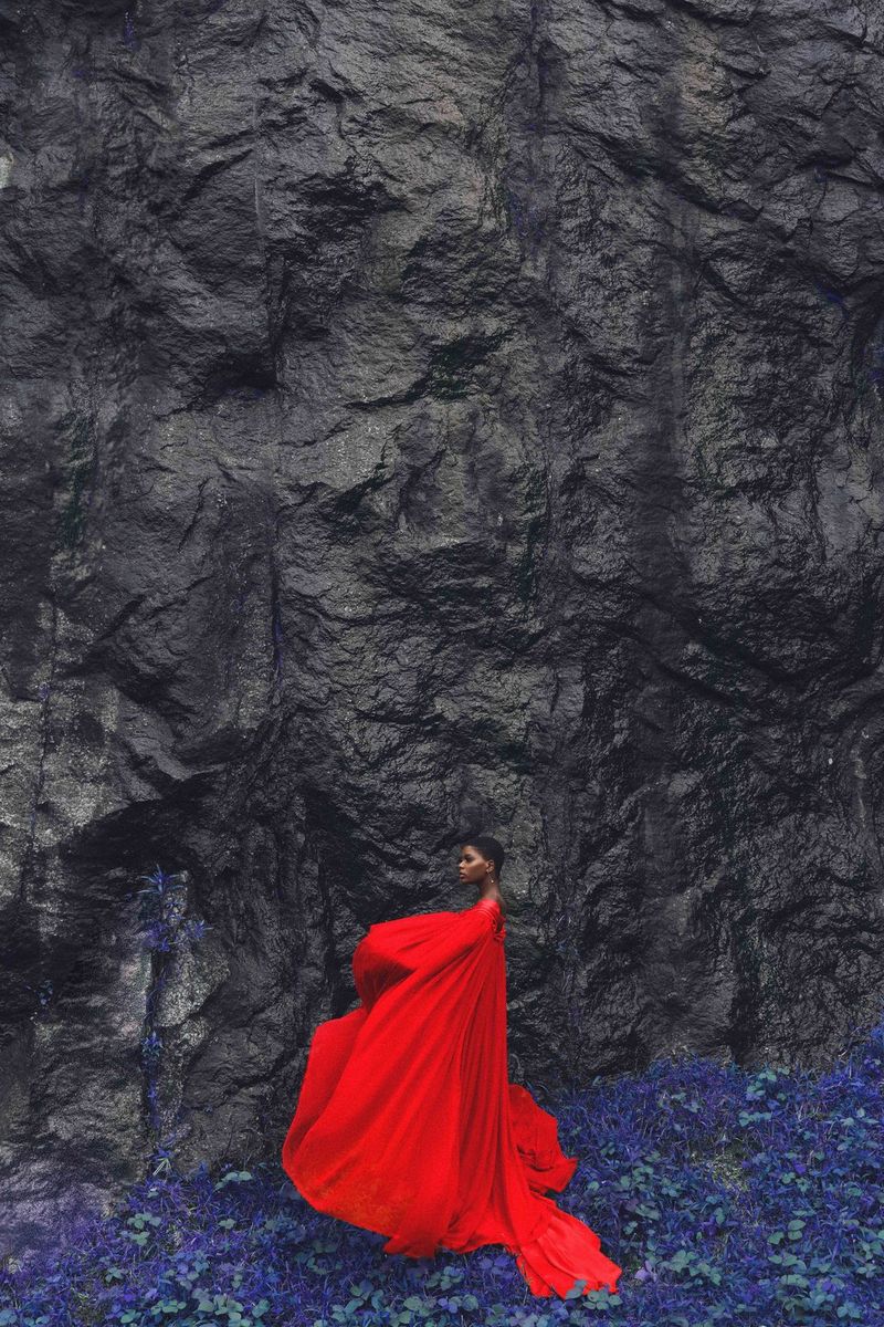 A woman in a striking red cape stands on a carpet of purple-blue flowers in front of dark grey rock.