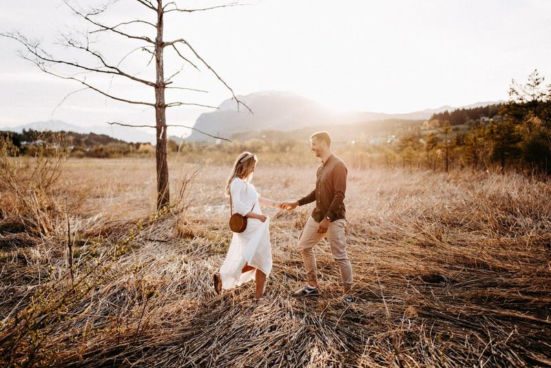 A couple smile and dance, holding hands, in a field of cut hay and a single bare tree.