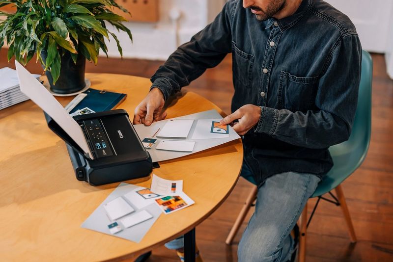 A man seated at a table examines the picture his Canon PIXMA TR150 printer has just produced.  