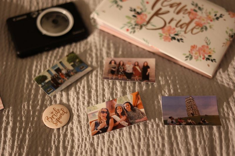 A Canon Zoemini S with a selection of prints and a badge, next to a photo album with Team Bride written on the front. 