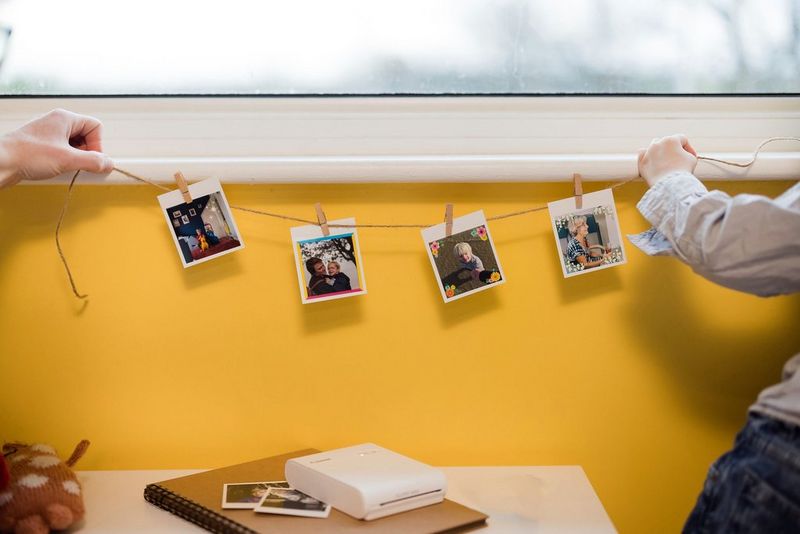 A parent and child hanging printed photos, clipped to a piece of string, from a window sill.