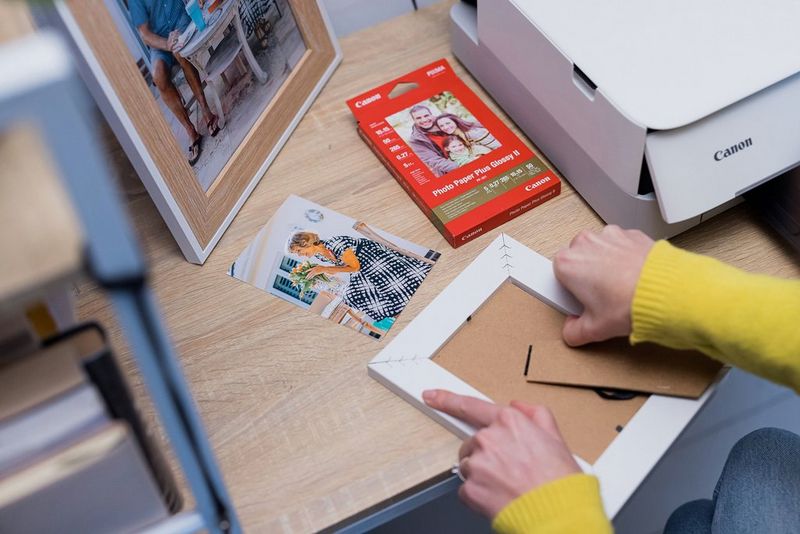 A woman removing the back of a picture frame. A Canon PIXMA TS5350a Series printer, a photo and a pack of glossy photo paper are on the desk in front of her.