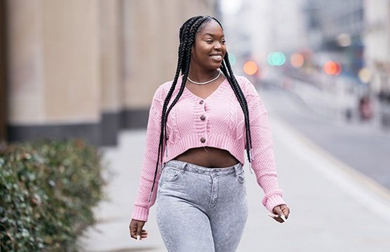 A portrait of a woman with long braids, wearing a pink cropped cardigan and grey jeans, walking along a city street.