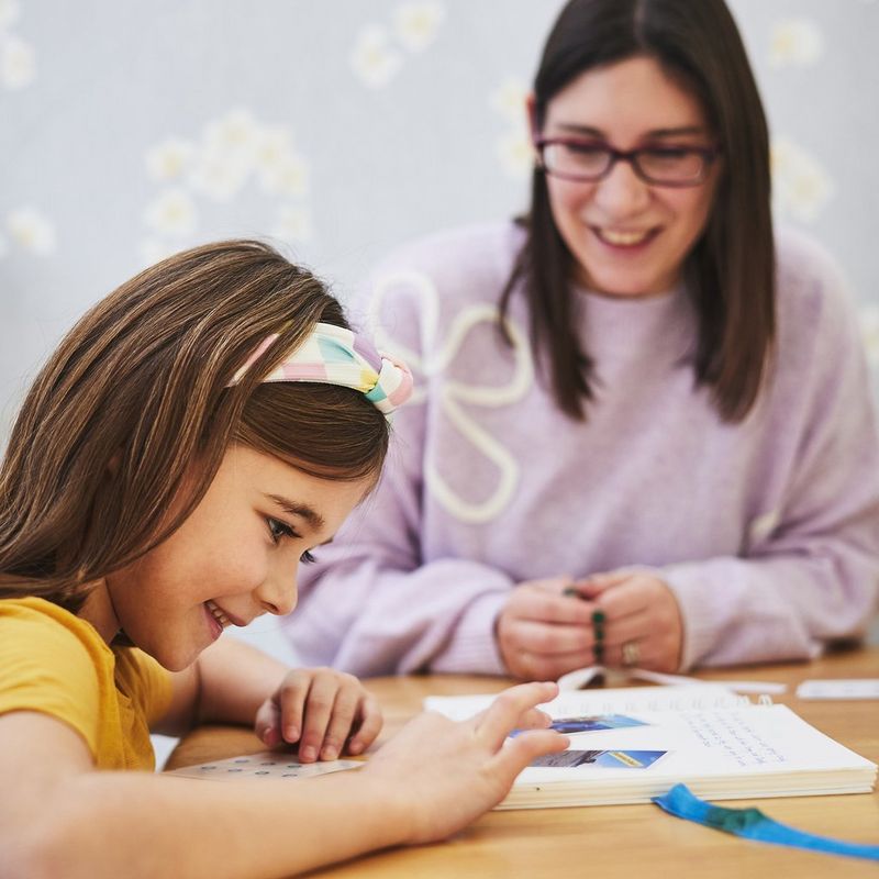 A smiling child holding a piece of coloured paper looks intently at the adult sat beside her.