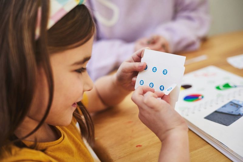 A smiling child peels off a small numbered sticker from a sheet of Canon sticker paper to put in the scrapbook in front of her.