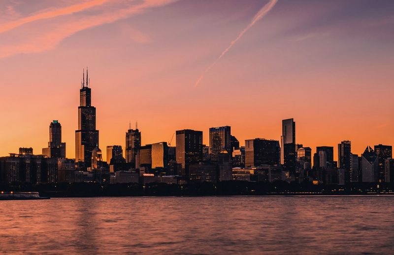 The Chicago skyline during golden hour, with water in front of the buildings. The entire scene is tinged with the orange glow of the horizon.