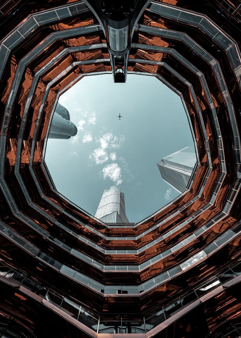 An image shot upwards through the hollow centre of a towering, circular structure. Rows of balconies form concentric circles up to the top of the structure, while a plane flies across the open sky.