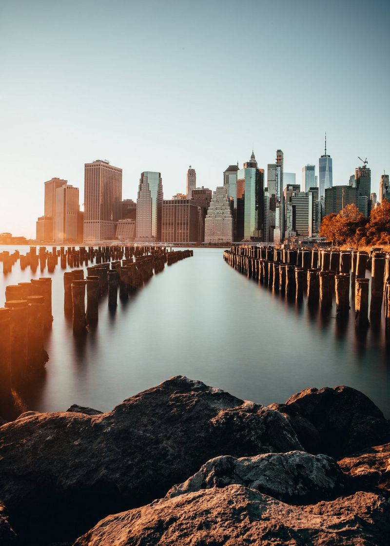 The New York City skyline seen from across a body of water, with rows of small pillars standing out of the water creating perspective lines towards the centre of the shot. A large, empty sky above the buildings is included in the image.