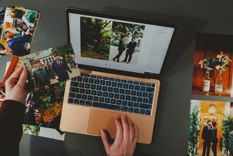 A user holds a photo print in one hand alongside a laptop, which is displaying photos of the same occasion. More photo prints are on the desk to each side of thelaptop.