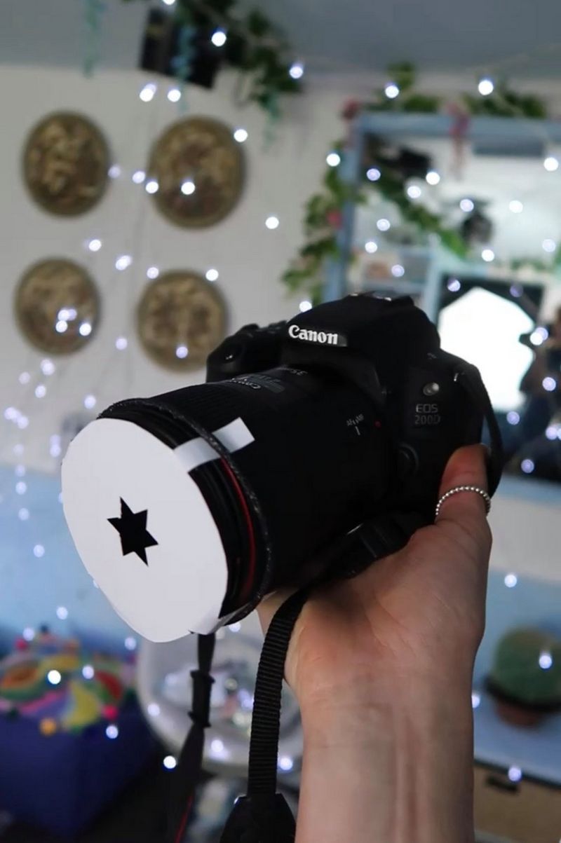 A hand holding up a Canon EOS 200D in front of a wall covered in fairy lights. A star-shaped paper cutout has been placed over the lens.