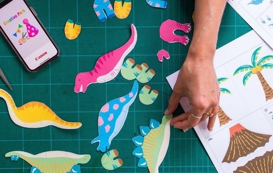 Various dinosaur templates printed from Canon Creative Park lie on a table, with a hand reaching to pick one up. 