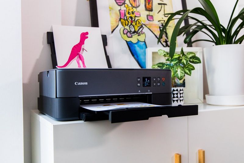 A dinosaur papercraft template emerges from a Canon PIXMA printer, with a completed model T-rex alongside pot plants to the side.