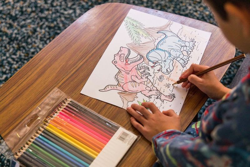 A child uses a brown pencil to colour in a drawing of dinosaurs.