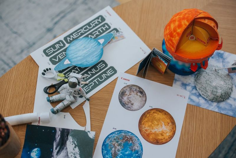 Egy papírból készült űrhajósfigura ül egy faasztalon, a Canon Creative Park alkalmazásból származó többi, űrtémájú papírsablonnal körülvéve.