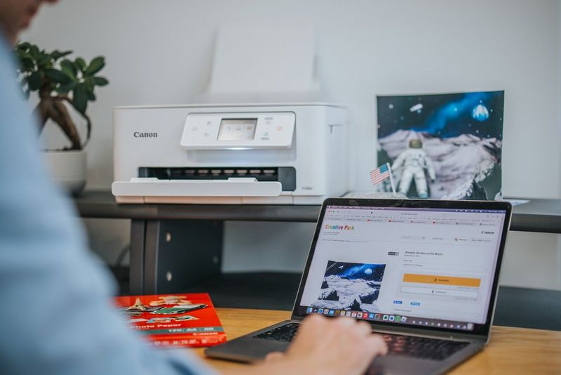 A person browses through the space-themed templates on Canon Creative Park on their laptop. Next to the laptop is a pack of Canon photo paper and behind it, a Canon PIXMA printer and an assembled papercraft astronaut sit on a shelf.